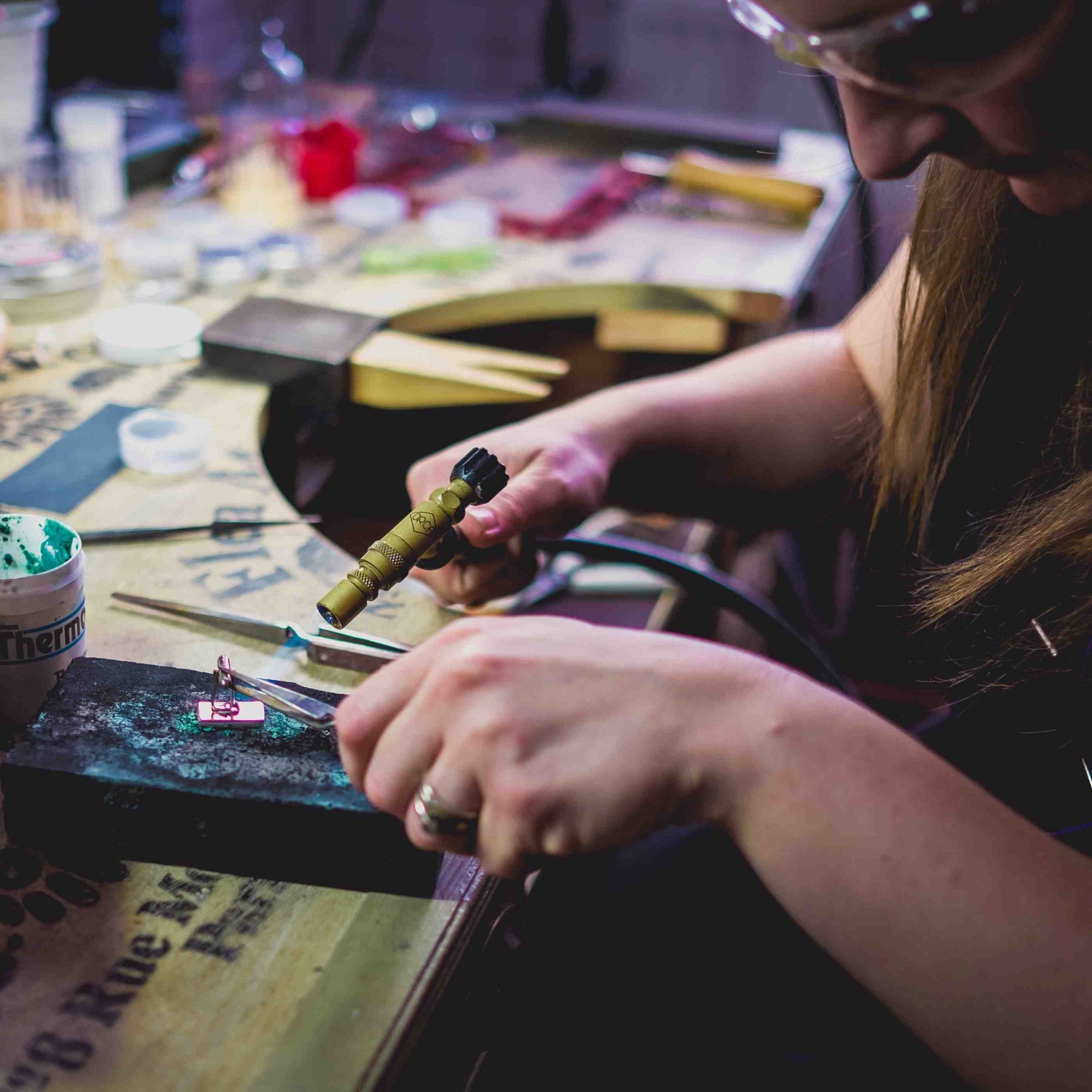 The Sophia Alexander Jewellery workshop.  Image shows Lucille Whiting soldering the back onto a gold footprint cufflink using a torch.  Tools can be seen scattered over the jewellery bench in the background.