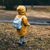 A toddler in a yellow raincoat with a small blue backpack walks outside, away from the camera on a rainy winter day.