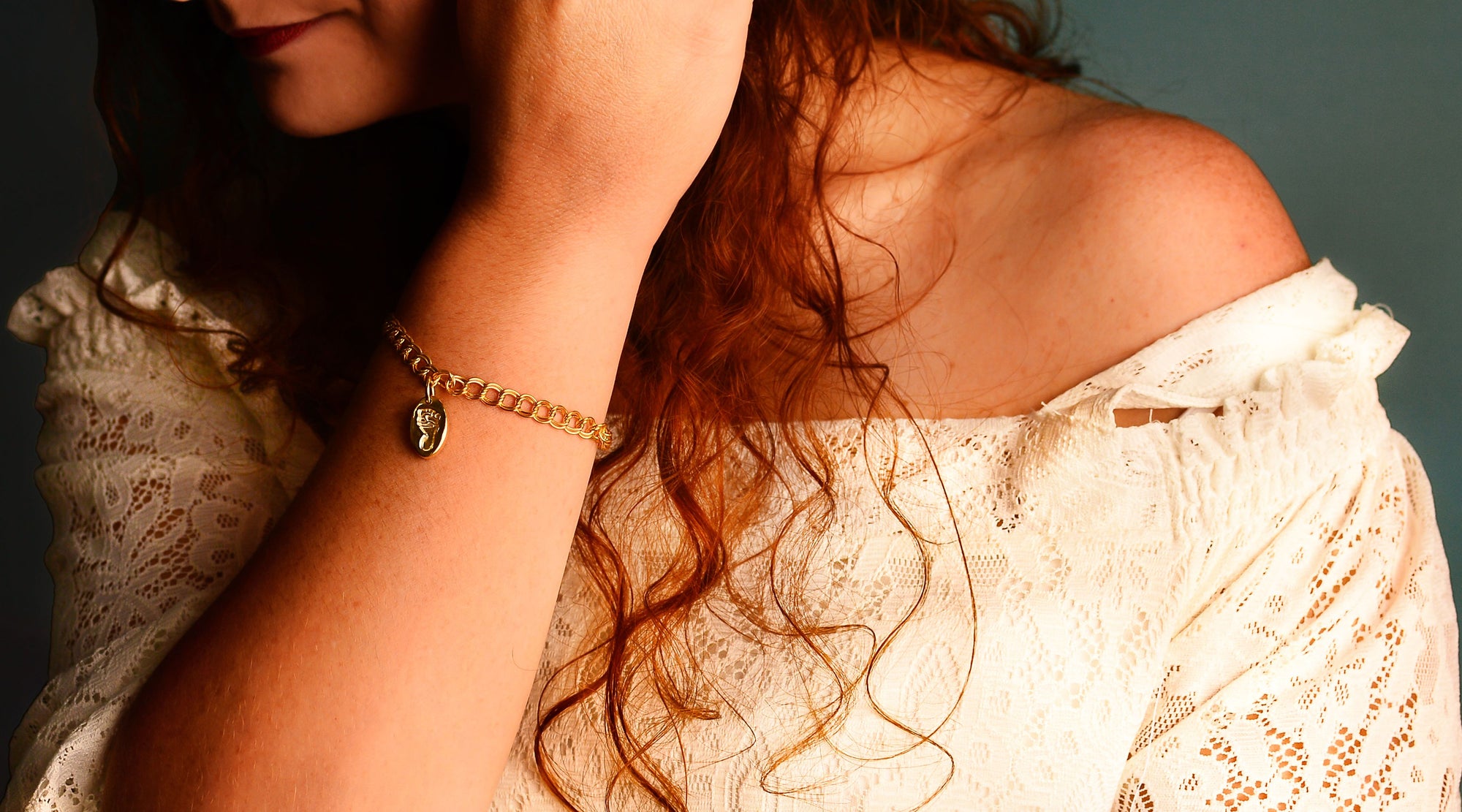 A solid gold baby footprint charm on a gold bracelet.  Worn by a model in a white lace top with a dark teal background behind her.