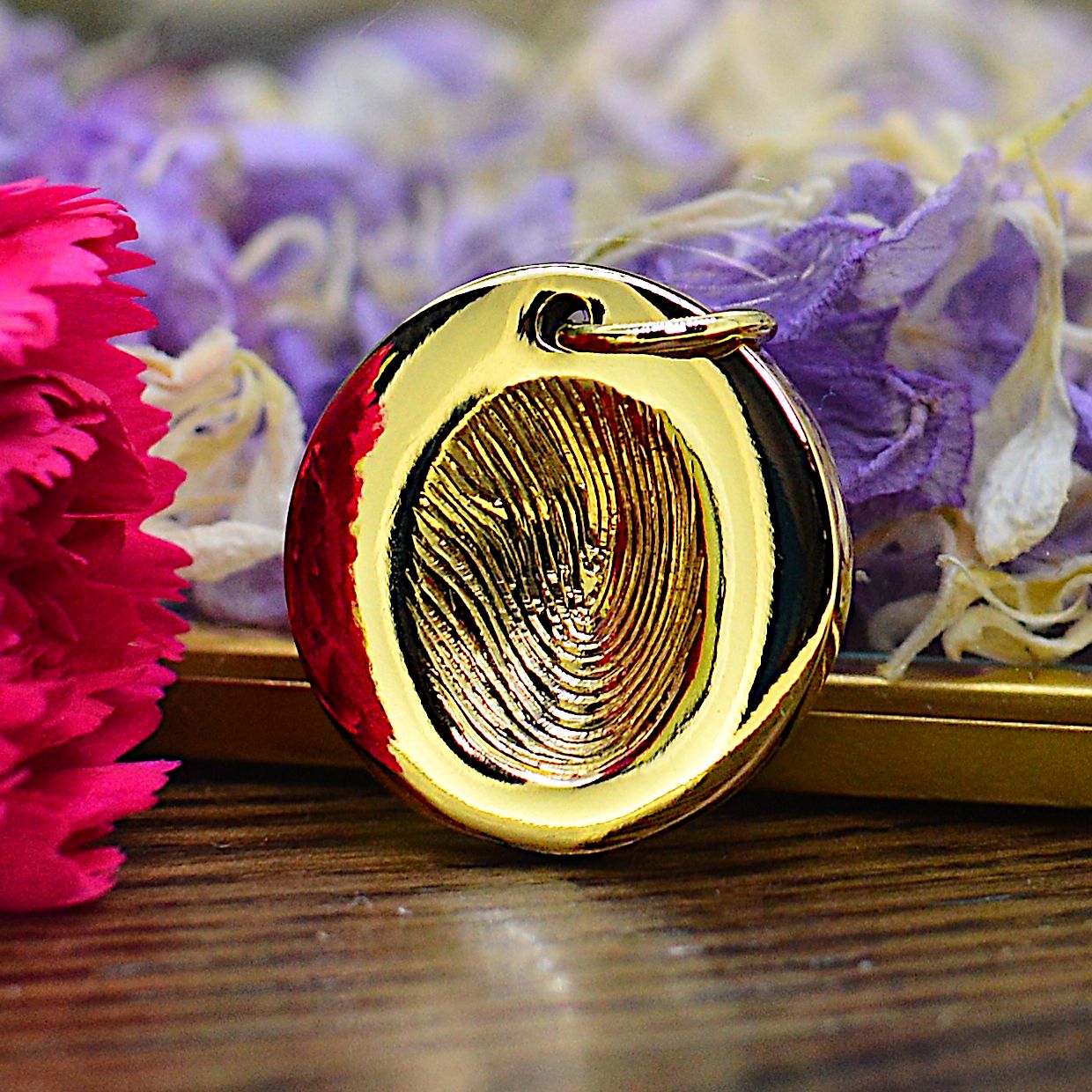 A solid gold disc-shaped necklace featuring an engraved fingerprint sits on a dark wooden background.  There is a ornate glass box behind it containing purple and white petals.  There is a pink carnation flower to the left side. 