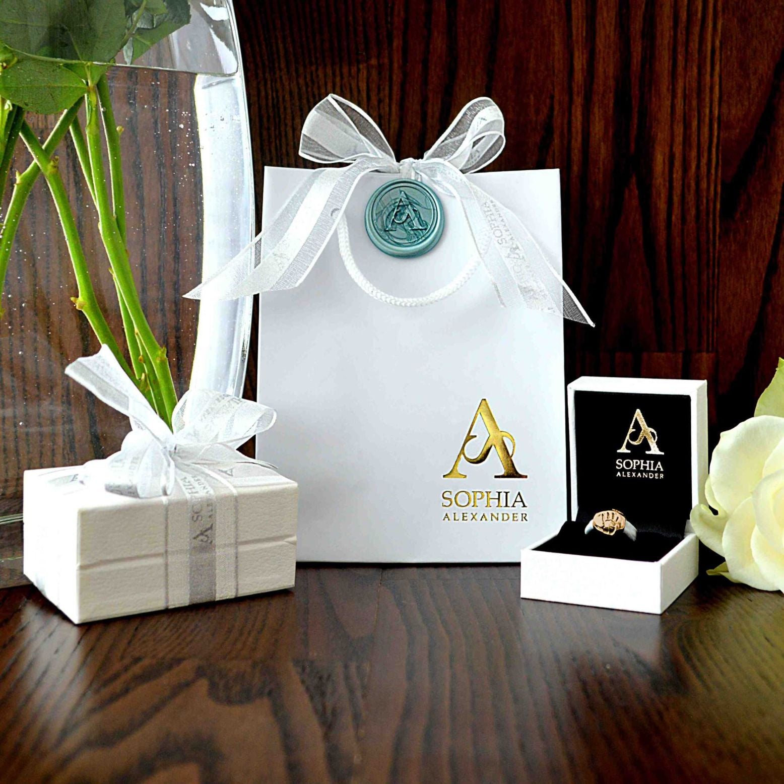 A Sophia Alexander Jewellery gift and two jewellery boxes sit on a dark wooden background.  A white rose is to the right side and a glass vase with rose stems is to the left side.  The bag and boxes are white with ribbons and a wax seal.  One jewellery box is open showing a gold charm bead that features a baby handprint.
