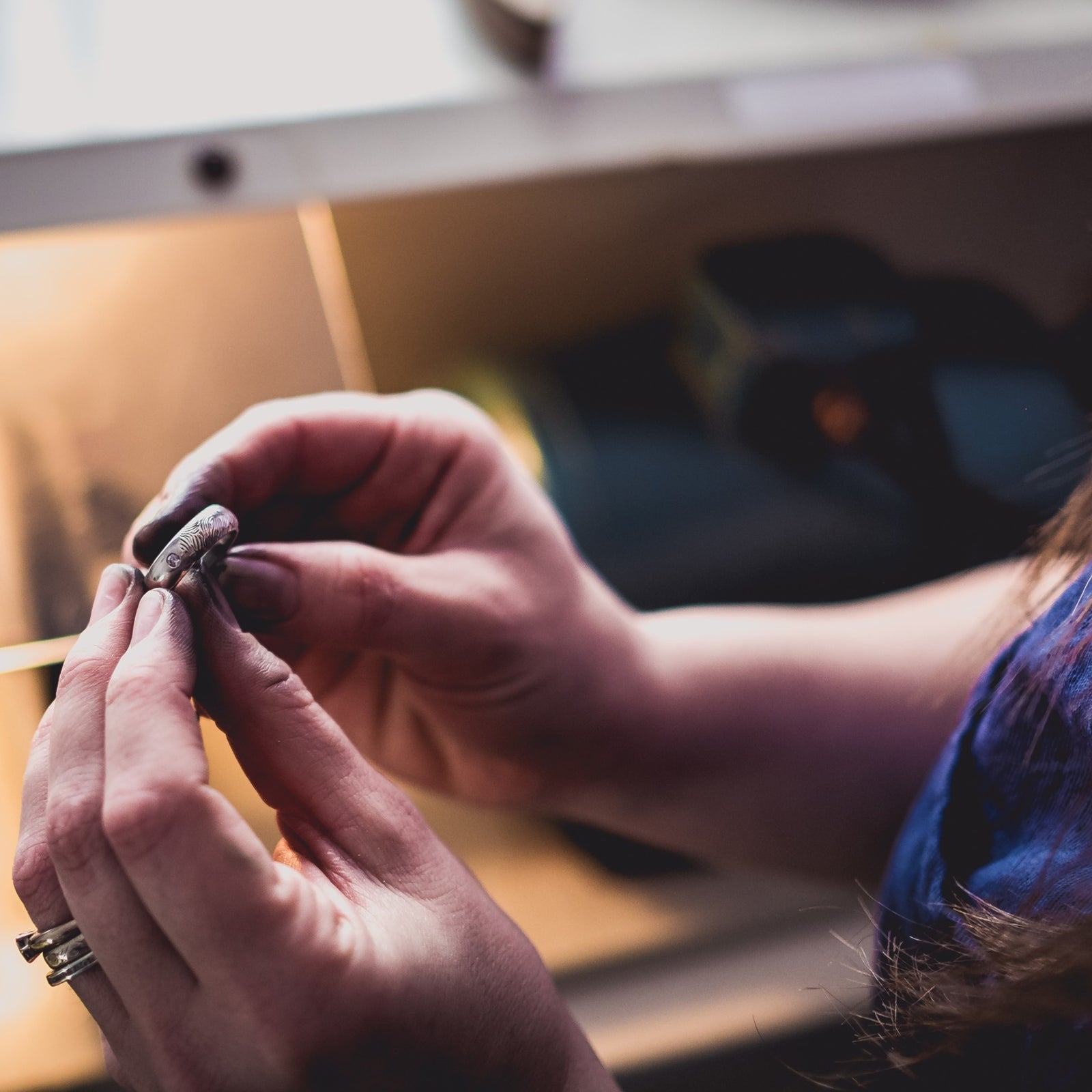 The Sophia Alexander workshop.  Lucille Whiting holds up a white gold wedding ring featuring a fingerprint and two diamonds.  Her two hands can be seen with a polishing machine in the background. 