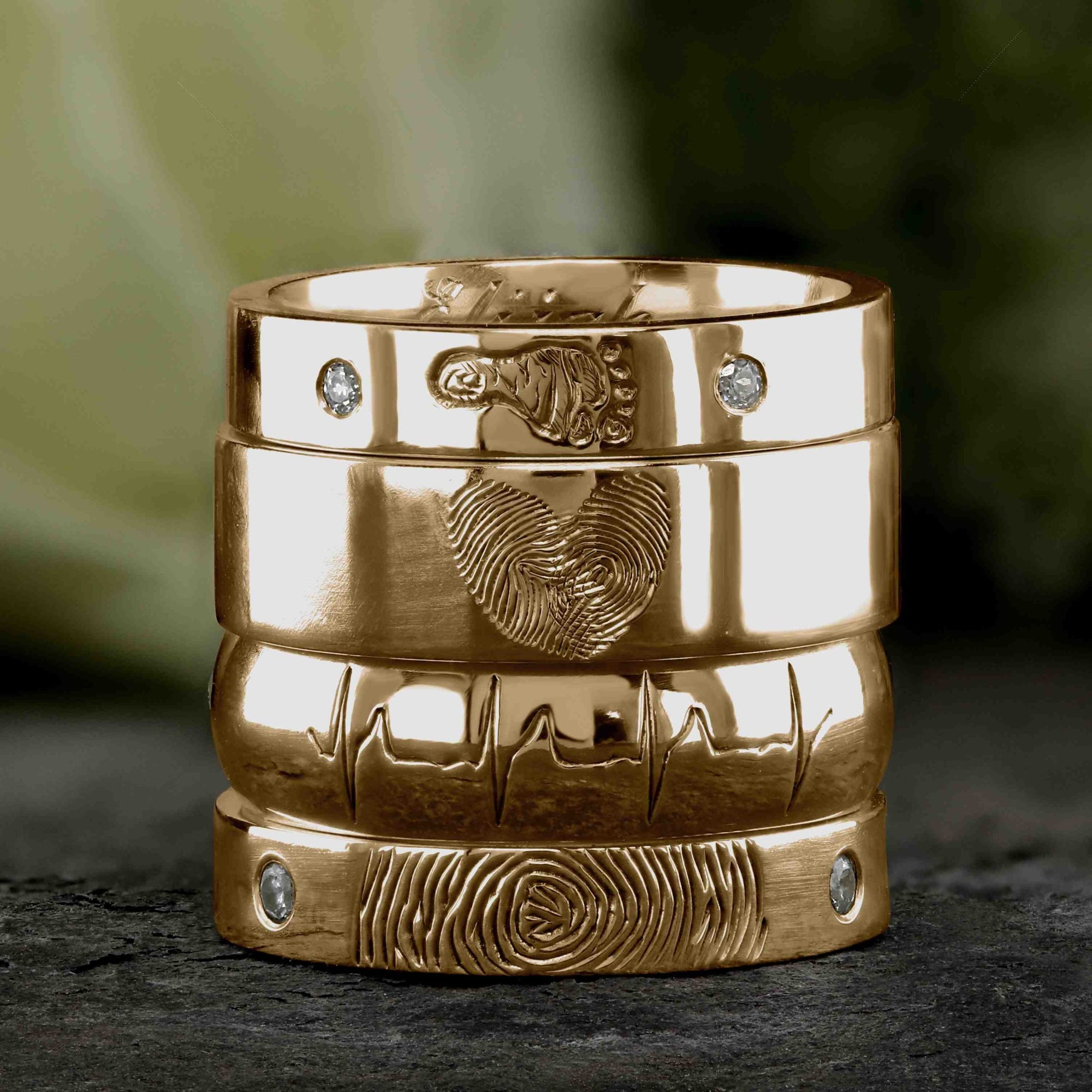 A stack of four yellow gold wedding rings, featuring engraved fingerprints, heartbeats, a baby footprint and diamonds.  They sit on a grey slate background with a white rose behind them.