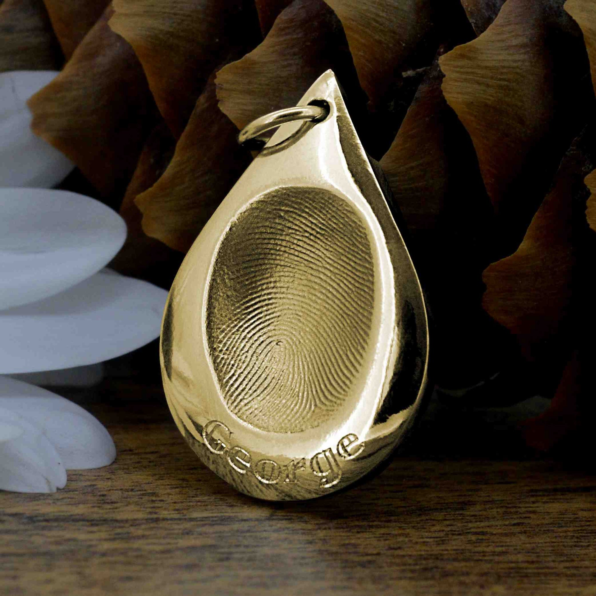 A solid gold teardrop-shaped necklace featuring a single adult fingerprint and the name George engraved underneath.  It sits on a dark wood background with a white flower and a pine cone behind it.