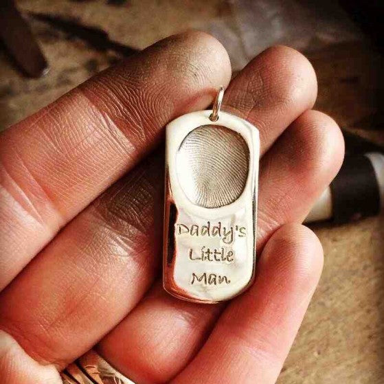 A solid silver dogtag with a single child's fingerprint and the engraved words "Daddy's Little Man".  It is being held on top of fingers that are covered with jewellery polish.  A workbench and tools can be seen in the background.