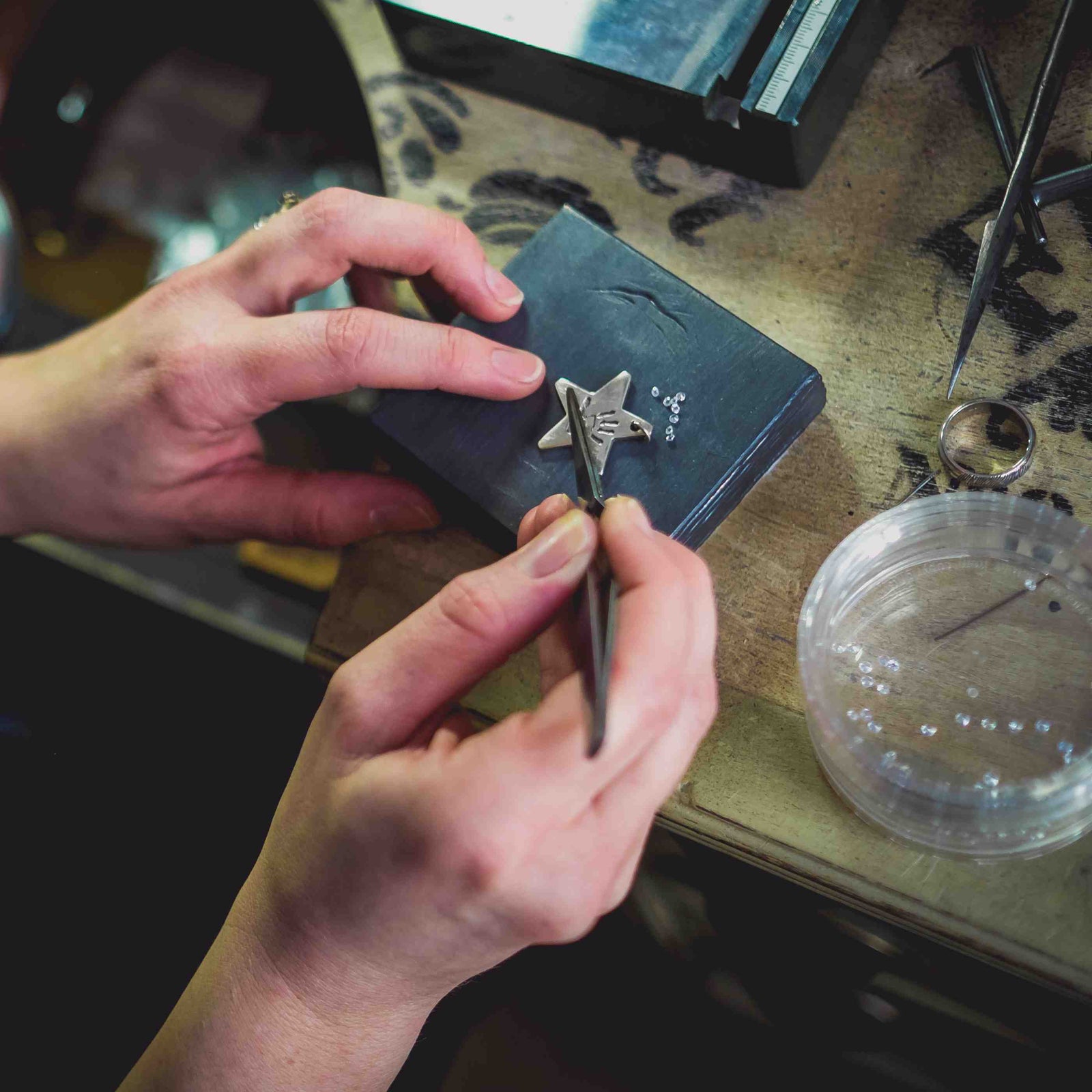 Two hands can be seen setting diamonds into a star-shaped necklace featuring a child's handprint.  It sits on a metal block.  A jewellery bench can be seen in the background with tools around it and a pot of diamonds off to the right side. 