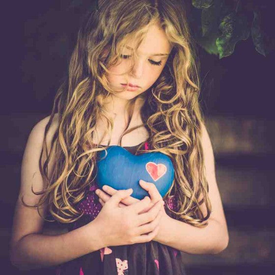 A young girl with ling curly blonde hair looks sadly down at the ground.  She holds a blue heart-shaped stone with a small red heart painted in the top right corner.  She stands in the shade with dark green leaves behind her.