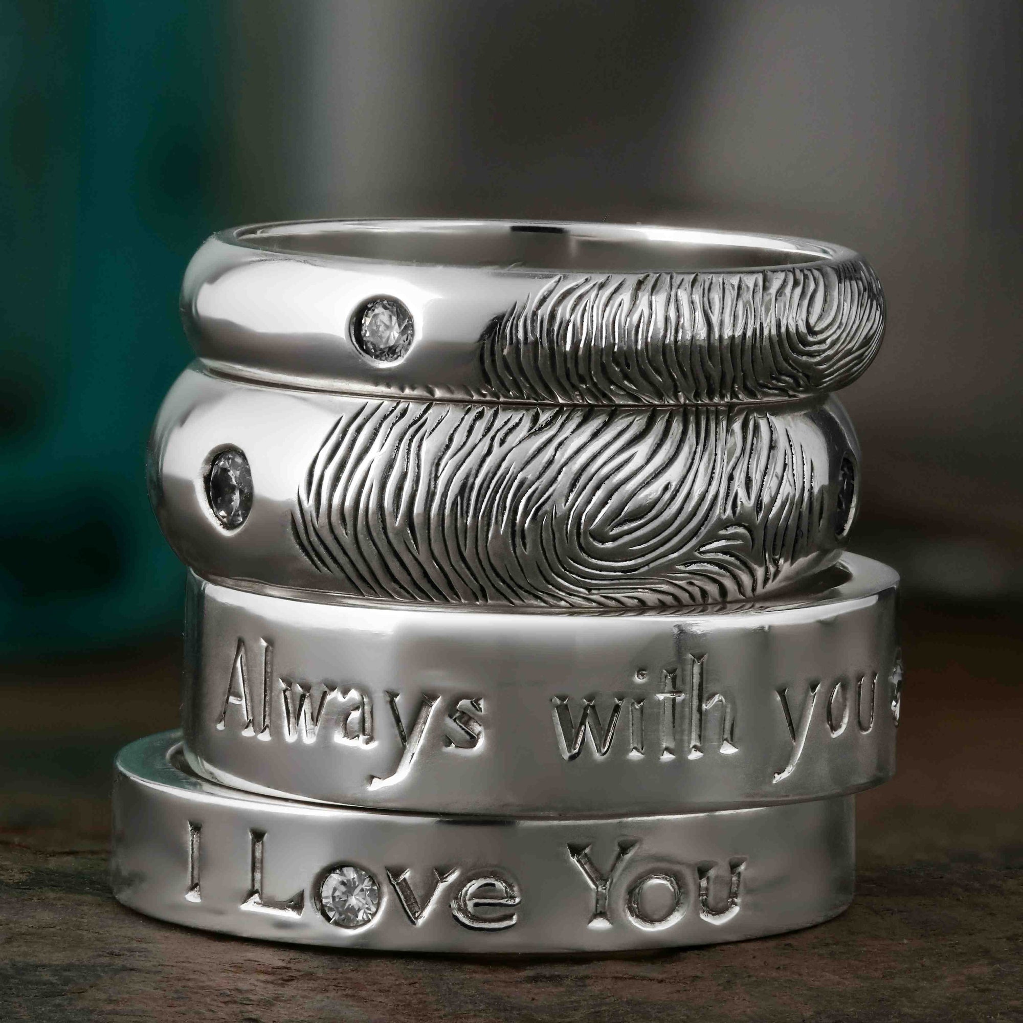 A stack of four platinum and white gold wedding rings, featuring engraved fingerprints, special words and diamonds.  One ring says "I love you" while another says "Always with you".  They sit on a dark wood background with teal silk behind them.