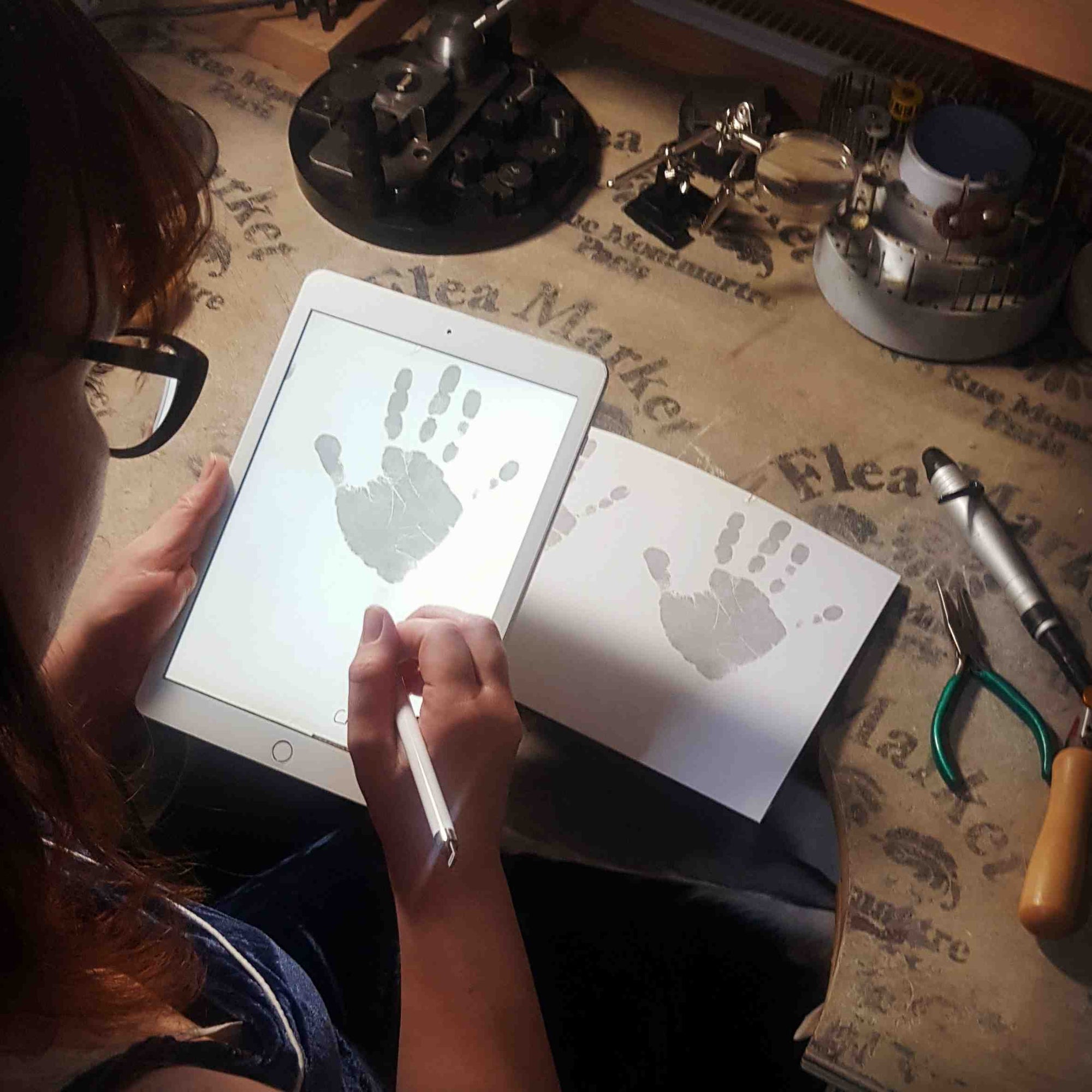 Lucille Whiting, owner at Sophia Alexander Jewellery sits at a jewellery bench with an Apple ipad.  The ipad screen shows a child's handprint.  She is editing it with an apple pencil.  On the bench are jewellery tools and a piece of paper showing the original handprint.