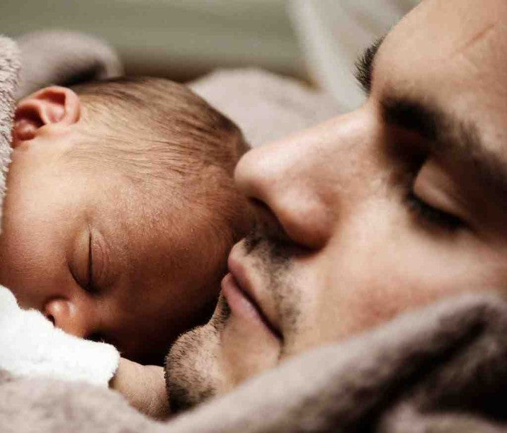 A father and newborn baby sleep with baby up against his Daddy's face.  They are cuddled together in a peddle coloured fleece blanket.
