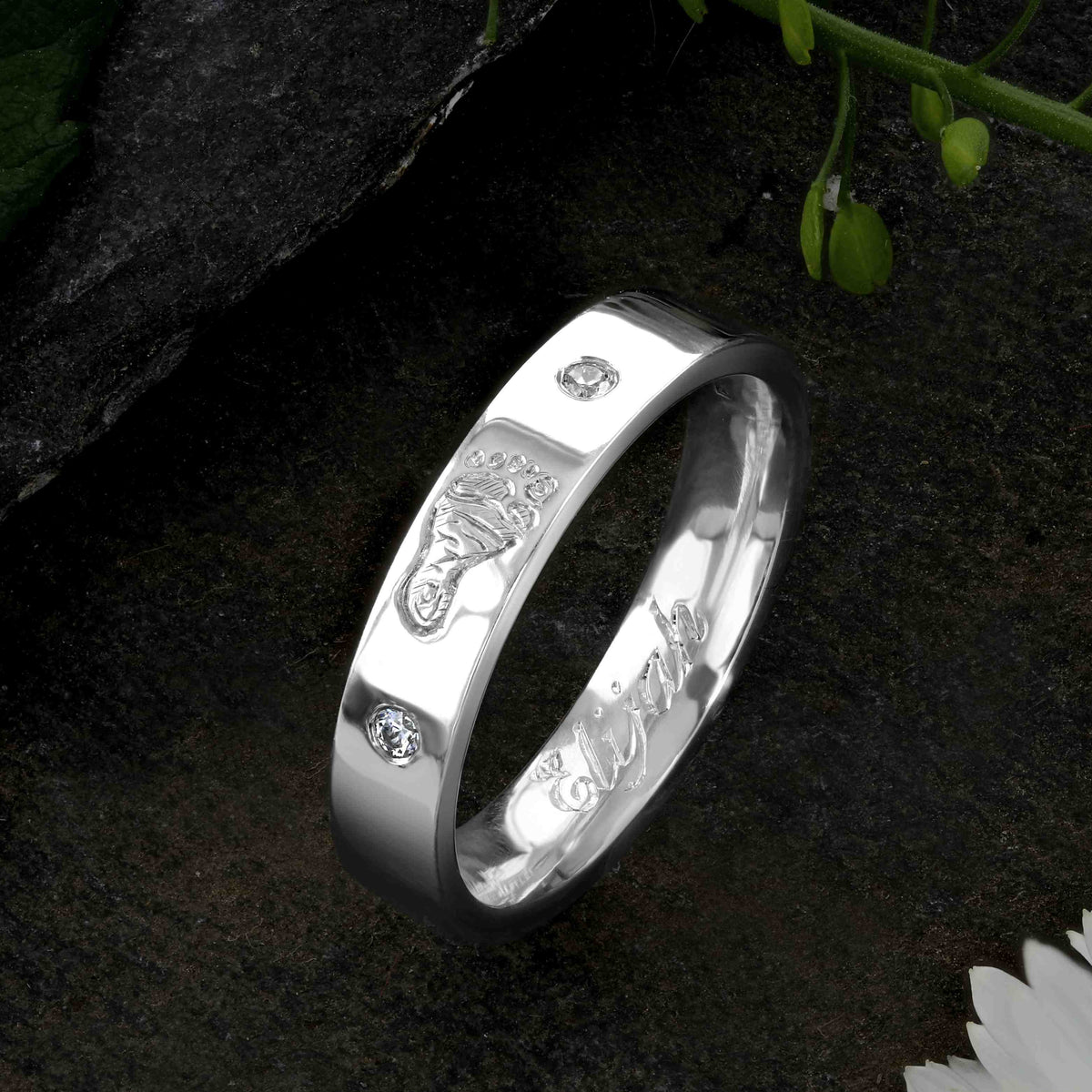 A White Gold Wedding Ring with an engraved baby footprint on the outer surface | Set with two diamonds that frame the footprint | Engraved with the name Elijah on the inside of the ring | Hand engraving | Ladies slim 4mm wide, comfort flat-court profile ring | Custom personalised wedding jewellery | Sophia Alexander Fingerprint Jewellery | Handmade in Suffolk UK